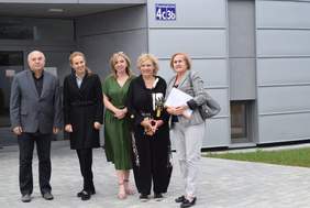 Pokaż zdjęcie: Na zewnatrz budynku stoi grupa 5 osób pozujących do zdjęcia, w tle szara elewacja budynku