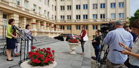 z lewej kobieta mówiąca do mikrofonów, fragment sylwetki mężczyzny na wózku inwalidzkim, kobieta tłumacząca na język migowy, z prawej strony grupa dziennikarzy, widać 5 osób, kamery