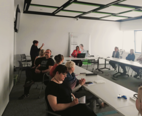 Uczestnicy spotkania, sziedzą przy stołąch. W tle widać tłumaczkę języka migowego i pracownika PFRON, prowadzącego spotkanie