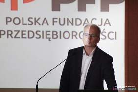 Pokaż zdjęcie: Przy mównicy stoi mężczyzna w garniturze i okularach.
