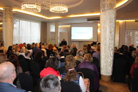 Pokaż zdjęcie: Zdjęcie sali konferencyjnej, robione od tyłu do przodu, widać plecy uczestników siedzących na krzesłach, w oddali ekran z prezentacją