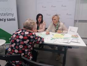 Pokaż zdjęcie: Rozmawiające osoby, tyłem starsza kobieta, za stołem dwie kobiety udzielające informacji. 