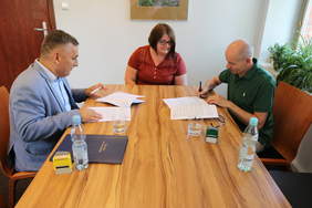 Pokaż zdjęcie: Przy stole siedzą trzy osoby, dwaj mężczyźni i kobieta po środku. W rękach trzymają długopisy i podpisują dokumenty. Są uśmiechnięci.