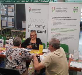 Pokaż zdjęcie: Kobieta siedzi za stolikiem na tle baneru PFRON oraz CIDON i udziela informacji starszemu małżeństwu, które przyszło do ZUS na Dzień Osób z Niepełnosprawnością. Na stoliku ułożone są materiały informacyjne promujące programy i zadania PFRON.