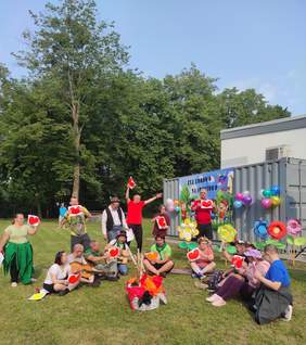 Pokaż zdjęcie: Grupa kilkunastu osób ubranych w barwne stroje siedzi lub stoi na trawniku przed budynkiem i metalowym kontenerem, na którym wisi baner promujący imprezę sportową. Dwie osoby mają gitarę, a większość trzyma w ręce czerwone serce na białej kartce.