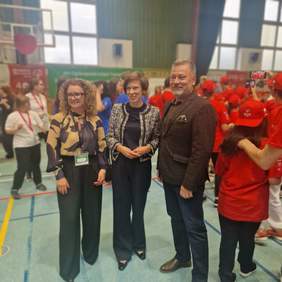 Pokaż zdjęcie: Dwie kobiety oraz jeden mężczyzna stoją w rzędzie i pozują do zdjęcia. W tle znajdują się sportowcy w mniejszych grupach, z których część także robi sobie wspólne zdjęcia.