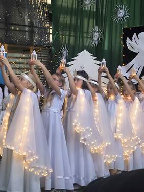 Pokaż zdjęcie: Osiem dziewczynek ubranych na biało jak aniołki, z białymi podświetlonymi skrzydłami, wznosi latarenki do góry.