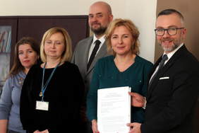 Pokaż zdjęcie: Stoją trzy kobiety i dwóch mężczyzn. Kobieta i mężczyzna prezentują podpisaną umowę. 