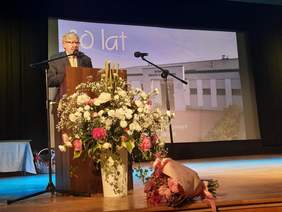 Pokaż zdjęcie: Na scenie kinoteatru w Skierniewicach stoi dyrektor Łódzkiego Oddziału PFRON. Stoi przy mównicy i przemawia do mikrofonu. Jest ubrany w garnitur. Na scenie znajdują sie dwa piękne i duże bukiety kolorowych kwiatów. 