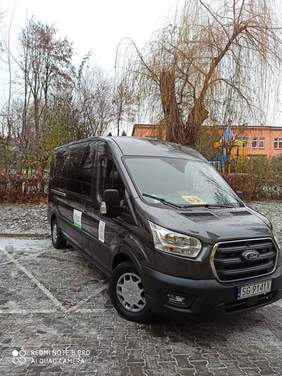 Pokaż zdjęcie: na pierwszym planie stoi nowy grafitowy bus z logo stowarzyszenie i PFRON w tle widać drzewa i budynek z placem zabaw