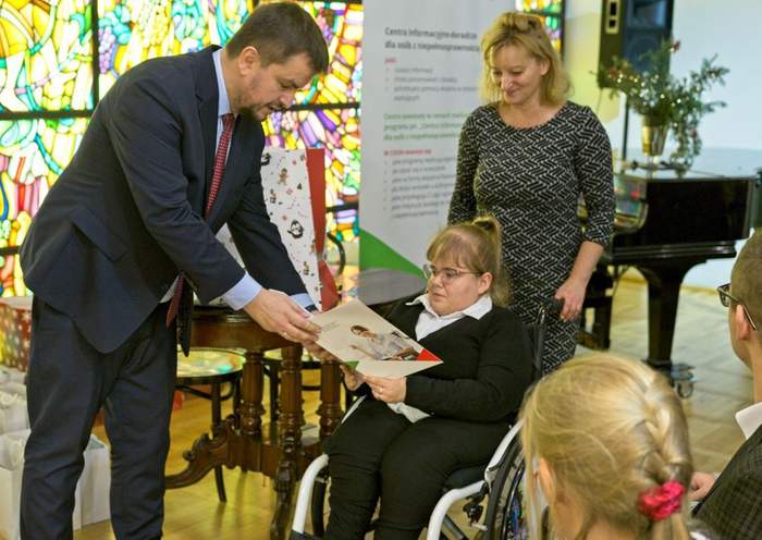 Pokaż zdjęcie: Dyrektor Oddziału wręcza nagrodę jednej z lauratek, która jest na wózku inwalidzkim, w tle jest rollup PFRON, witraże zdobiące Muzeum