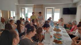 Pokaż zdjęcie: Dwie kobiety stoją, pozostałe osoby siedzą przy stole, słuchając wypowiedzi kobiet. Trwa zakończenie roku szkolnego.