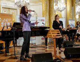 Pokaż zdjęcie: Artysta śpiewa trzymajac w ręku mikrofon. W tle stoi fortepian. Po prawej stronie kilkaosób gra na skrzypcach.