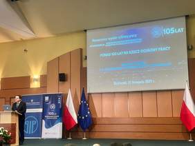 Pokaż zdjęcie: Mężczyzna w garniturze przemawia na podium podczas konferencji. W tle widoczny jest  slajd z tytułem Ponad 100 lat na rzecz ochrony Pracy, Dr Karol Łapiński, p.o. Okręgowego Inspektora Pracy w Białymstoku, Białystok, 21 listopada 2024 r., po bokach polska i unijna flaga oraz banery informacyjne z logo Państwowej Inspekcji Pracy. 