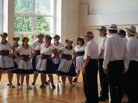 na wprost na tle dużego okna w sali grupa kobiet w stojach ludowych, trzymaja się pod boki, tańczą, z prawej strony mężczyźni