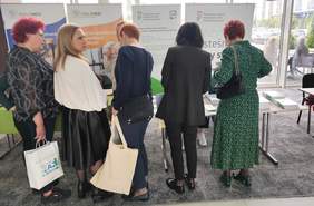 Pokaż zdjęcie: Pięć kobiet w średnim wieku nachyla się nad stolikiem. Na stoliku położone są ulotki i materiały informacyjne. Za stolikiem widoczny rollup z logotypem PFRON