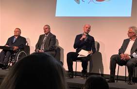 Pokaż zdjęcie: mężczyźni uczestniczący w debacie siedzą na krzesłach ustawionych na scenie. nad nimi na ścianie wyświetlana jest prezentacja.