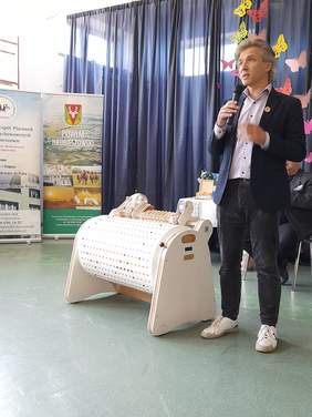 Pokaż zdjęcie: Prelegent opowiada o urządzeniu musicon, które stoi koło niego, jest to urządzenie w jasnym kolorze, wydłużony kształt walca z którego wychodzą różne wypustki emitujące różne dźwięki.