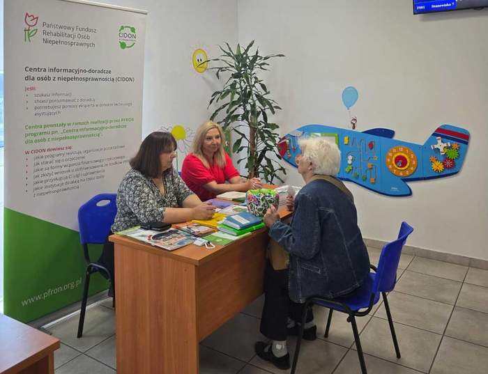 Pokaż zdjęcie: Przy stoliku na którym leżą ulotki siedzą trzy kobiety i rozmawiają. W tle rollup z logo funduszu.