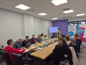 Pokaż zdjęcie: Stół konferencyjny przy którym siedzą uczestnicy spotkania, z przodu stoi prelegentka w beżowej marynarce.