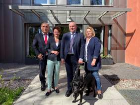 Pokaż zdjęcie: Na zdjęciu stoją ubrani na galowo ludzie na tle wejścia do budynku. Jeden z mężczyzn trzyma w ręku uprząż do psa asystującego. Wejście wykonana z ciemnej stali  z dużym, i szklanymi oknami i szklanym, zadaszeniem.