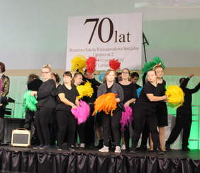 Pokaż zdjęcie: Na scenie dzieci przebrane w czarne ubrania tańcza w rytm i do melodii, machając kolorowymi pomponami z bibuły