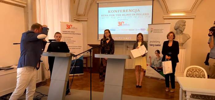 Pokaż zdjęcie: Cztery kobiety stoją na scenie przed momentem wręczenia nagród w Konkursie Idol 2022. Po lewej stronie stoi mężczyzna, który robi zdjęcia. W tle widoczne są banery Fundacji Szansa dla Niewidomych oraz Państwowego Funduszu Rehabilitacji Osób Niepełnosprawnych.