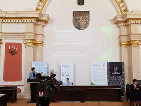 Pokaż zdjęcie: Na mównicy stoi kobieta na tle baneru OWiT w Kaliszu. Na środku ustawiona jest kamera Telewizji Kalisz. Na sali widoczne są banery: PFRON, CIDON oraz Miasta Kalisza. Na ścianie wyświetlana jest prezentacja. W sali siedzi też tłumacz języka migowego.