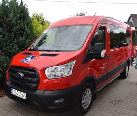 Pokaż zdjęcie: Czerwony bus z naklejką informującą, że pojazd przystosowany jest do przewozu osób z niepełnosprawnością. W bocznej szybie samochodu odbija się jak w lusterku grupa kilku zgromadzonych w pobliżu osób. W tle drzewa i krzewy oraz fragment budynku.