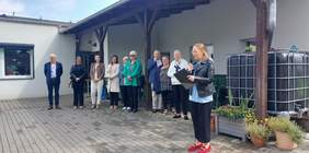 Pokaż zdjęcie: Grupa dziesięciu osób stoi przed zadaszonym wejściem do budynku. Kobieta po prawej stronie trzyma w dłoniach mikrofon oraz podkładkę na dokumenty.