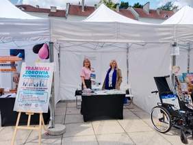 Pokaż zdjęcie: Dwie kobiety stoją na stoisku Państwowego Funduszu Rehabilitacji Osób Niepełnosprawnych. Pozują do zdjęcia. Uśmiechają się. Na stole leżą różne informatory, broszury, ulotki. 