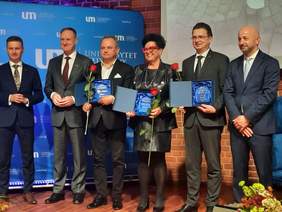 Pokaż zdjęcie: W sali stoi kobieta w otoczeniu pięciu mężczyzn. Pani i dwóch panów trzymają w dłoniach statuetki i czerwone róże. Wszyscy są elegancko ubrani. Pozują do zdjęcia. 