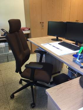 Pokaż zdjęcie: na pierwszym planie duży brązowy fotel biurowy przed nim widać fragment biurka z dwoma monitorami i klawiaturą w tle stoją wysokie zamykane szafy