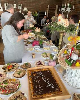 Pokaż zdjęcie: Wokół wielkanocnych stołów zgromadzeni są ludzie. Częstują się światecznymi potrawami. Stoły są pięknie udekorowane wielkanocnymi ozdobami. 