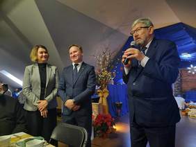 Pokaż zdjęcie: Przy stole stoi mężczyzna w garniturze, który przemawia do mikrofonu. Składa świąteczne życzenia. Obok niego stoi kobieta i drugi mężczyzna, którzy się uśmiechają. 