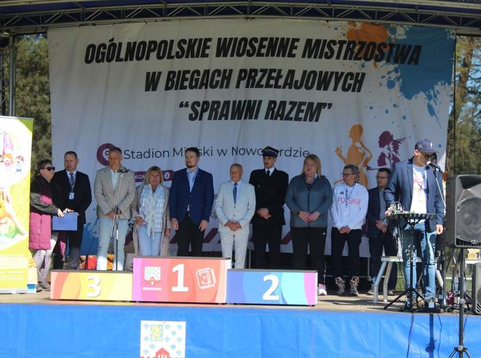 Pokaż zdjęcie: Grupa osób stoi na scenie pod zadaszeniem podczas powitania gości i rozpoczęcia biegu. W tle znajduje się baner z napisem: „Ogólnopolskie Wiosenne Mistrzostwa w Biegach Przełajowych ‘Sprawni Razem’”. Scena udekorowana jest również logo miasta Nowogard i sponsorów.