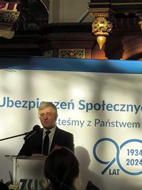 Pokaż zdjęcie: Na zdjęciu znajduje się mężczyzna w średnim wieku, elegancko ubrany w ciemny garnitur, białą koszulę oraz krawat. Przemawia do mikrofonu, stojąc za przezroczystą mównicą. W tle widoczny jest baner z napisem "Zakład Ubezpieczeń Społecznych" oraz hasłem związanym z obchodami 90-lecia instytucji, oznaczonymi datami "1934–2024". W górnej części kadru można dostrzec elementy dekoracyjne wnętrza, takie jak ozdobne drewniane detale oraz fragment modelu statku, co sugeruje historyczną scenerię miejsca wydarzenia. Atmosfera jest oficjalna i poważna, typowa dla uroczystych spotkań i jubileuszów.