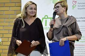 Pokaż zdjęcie: Dwie kobiety stoją na z mikrofonem i kartkami w dłoniach podczas otwarcia Konferencji „Technologie asystujące wspierające wczesne wspomaganie rozwoju dziecka”. W tle widoczny baner z logotypami organizatorów. 