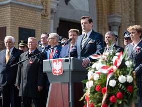 Pokaż zdjęcie: na zdjęciu mównica na której przemawia Wojewodoa Łodzki obok i w tle stoją przedstawiciele władz samorządowych i rządowych oraz inni uczestnicy wydarzenia