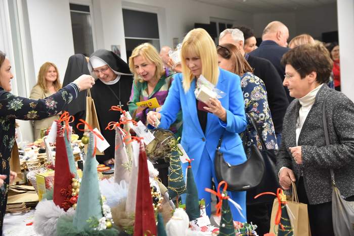 Pokaż zdjęcie: Przy stoliku stoi duża grupa osób kupujących różne rzeczy stojące na stoliku, takie jak, ozdoby  świąteczne, mikołaje, biżuteria, ceramika, trwa kiermasz charytatywny