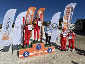 Trzy osoby, narciarze na podium, po prawej jeszcze dwie, słoneczny dzień, za nimi banery reklamowe