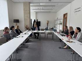 Pokaż zdjęcie: Kilka osób siedzi przy stołach w sali konferencyjnej, prowadząc spotkanie. Część notuje, część słucha. Na ścianie widoczny cytat: „Rób rzeczy wielkich, w wielki sposób”. W tle kanapy, lampy i regał z filiżankami.