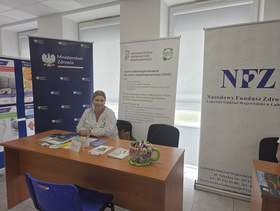 Stoisko informacyjne PFRON podczas wydarzenia. Przy biurku siedzi pracownik, w tle widoczne są roll-upy PFRON, CIDON, NFZ i Ministerstwa Zdrowia oraz materiały informacyjne.