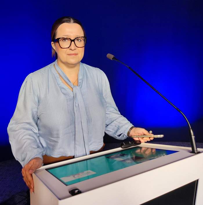 Pokaż zdjęcie: Przy mównicy stoi kobieta w okularach, ubrana w niebieska bluzkę.