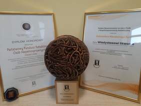 Pokaż zdjęcie: Na stoliku stoi statuetka w kształcie kuli. Po jej prawej i lewej stronie znajdują się dwa dyplomy. 