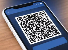 Pokaż zdjęcie: Smartfon w jasnym etui leżący na drewnianym blacie. Na ekranie aplikacja w niebieskiej kolorystyce z dużym, czarno-białym kodem QR wyświetlonym centralnie, widok pod lekkim kątem.