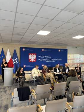 Pokaż zdjęcie: Przy stole siedzi kilka osób. Po lewej stronie kobieta stoi przy mównicy. W tle widoczne są flagi Polski, Unii Europejskiej i NATO oraz baner z napisem „Wojewoda Warmińsko-Mazurski”.