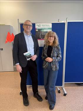 Pokaż zdjęcie: Dwie osoby stoją obok siebie w pomieszczeniu biurowym na tle niebieskich tablic informacyjnych. Mężczyzna w marynarce i jasnej koszuli trzyma w ręku ulotkę, kobieta ubrana jest w wzorzysty żakiet i jeansy. Za nimi widoczny jest plakat z napisem „OWiT – Ośrodek Wsparcia i Testów w Olsztynie”.