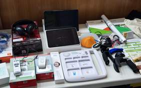 Pokaż zdjęcie: Zbliżenie na asortyment urządzeń i akcesoriów pomocniczych dla osób z niepełnosprawnościami, leżących na białym stole. Wśród nich jest duży biały komunikator z ekranem dotykowym i obrazkami (QuickTalker), tablety, słuchawki, piloty zdalnego sterowania oraz broszury informacyjne.