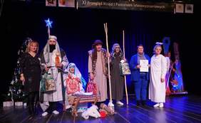 Pokaż zdjęcie: Grupa uczestników XI Przeglądu Kolęd i Pastorałek Wielowyznaniowych stoi na scenie Domu Ludowego w Supraślu. Osoby ubrane są w stroje nawiązujące do jasełek – pasterzy, anioła i postaci biblijnych. Na scenie widoczne są świąteczne dekoracje, choinka oraz rekwizyty bożonarodzeniowe.